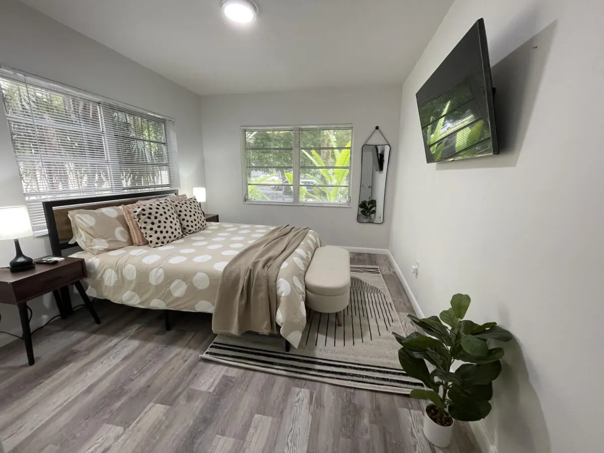 A bright, inviting bedroom featuring a bed with cheerful polka dot bedding, a wall-mounted smart TV, a lush green plant in the corner, and a large window streaming in natural Miami sunlight. The space feels cozy, serene, and perfectly suited for relaxation or unwinding with your favorite show.