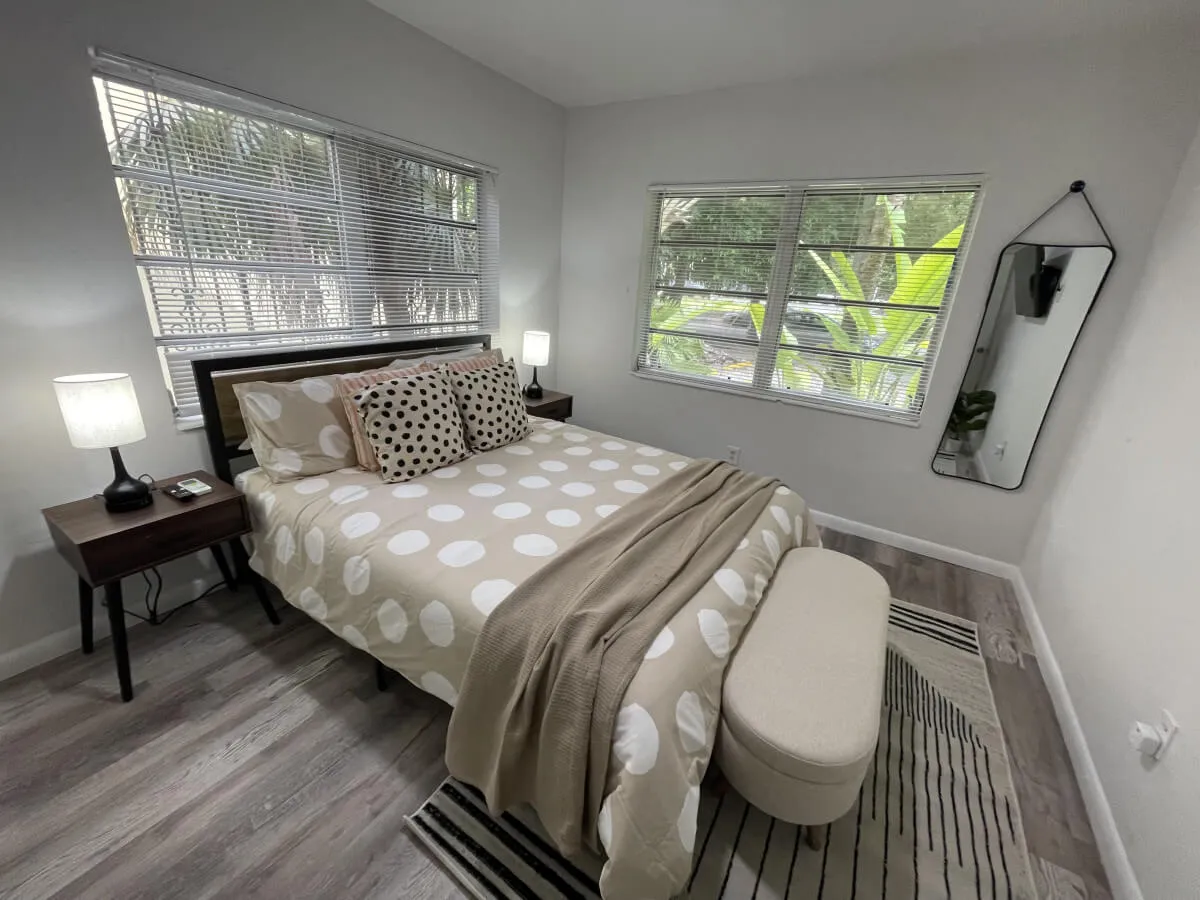 This image features a serene bedroom with a plush queen bed dressed in polka dot bedding, flanked by bedside lamps. Large windows offer a view of greenery, filling the room with natural light, while a full-length mirror adds both function and style. The mood is tranquil and inviting—ideal for unwinding after a day out.