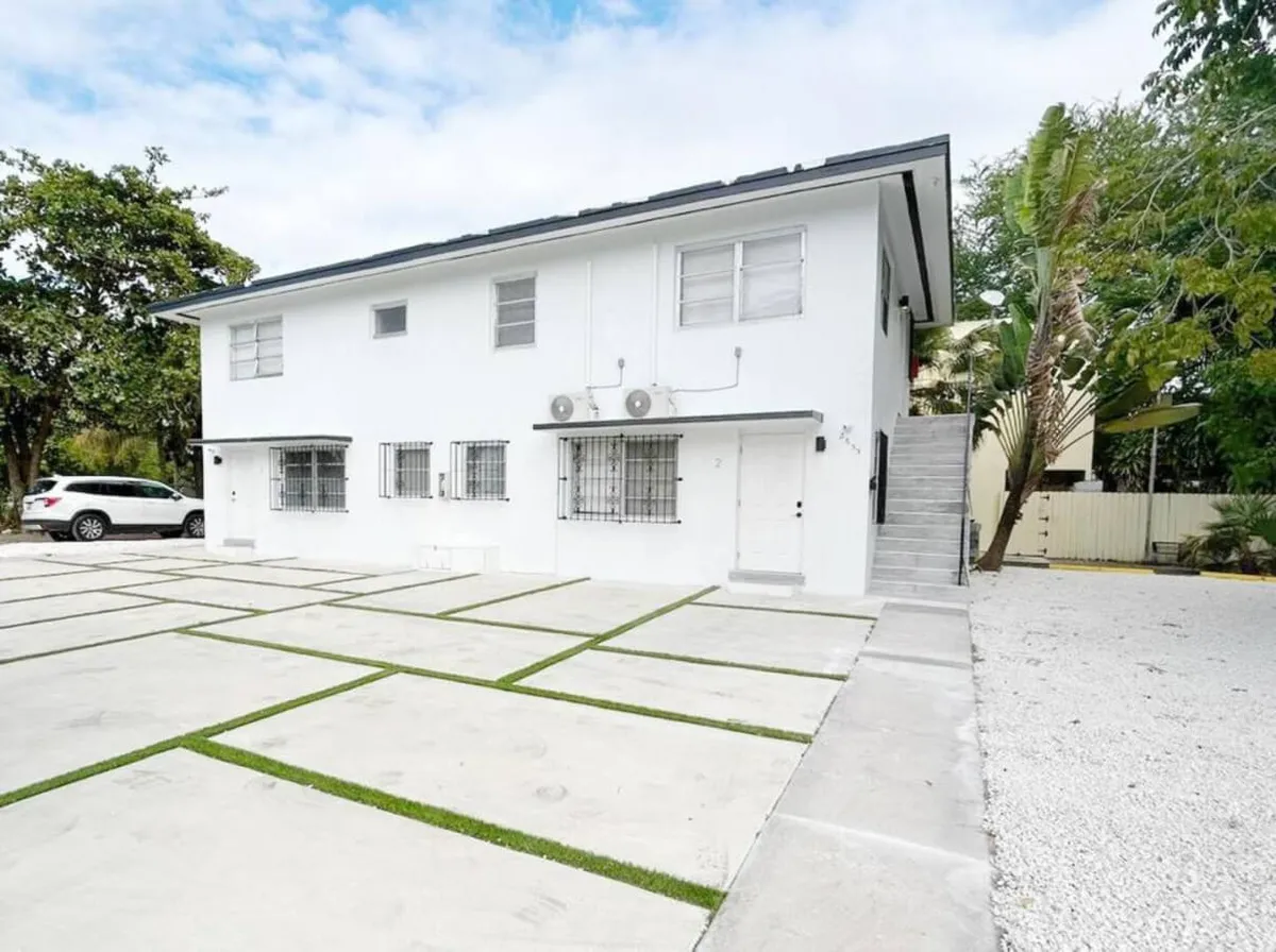 This exterior photo features the white building facade surrounded by green grass, an outdoor stairway, and mature trees. The parking area is visible, giving a sense of the property's welcoming exterior and the convenience of on-site parking. The community atmosphere is suggested by social areas suitable for gatherings.