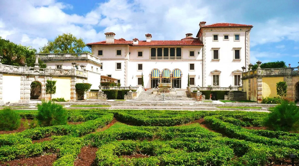 This image captures the impressive exterior of an ornate mansion surrounded by manicured gardens, elegant stone pathways, and a central fountain. The lush outdoor environment and architectural details create a sense of arrival and sophistication, suggesting a prime location with easy access to attractive local areas.