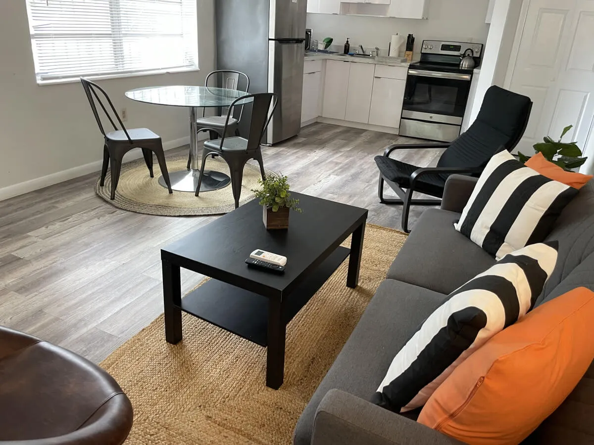 This inviting open-plan space connects a cozy living area with a glass dining table adjacent to visible kitchen appliances. The colorful cushions and bright ambiance make it perfect for gathering, dining, or relaxing with coffee.