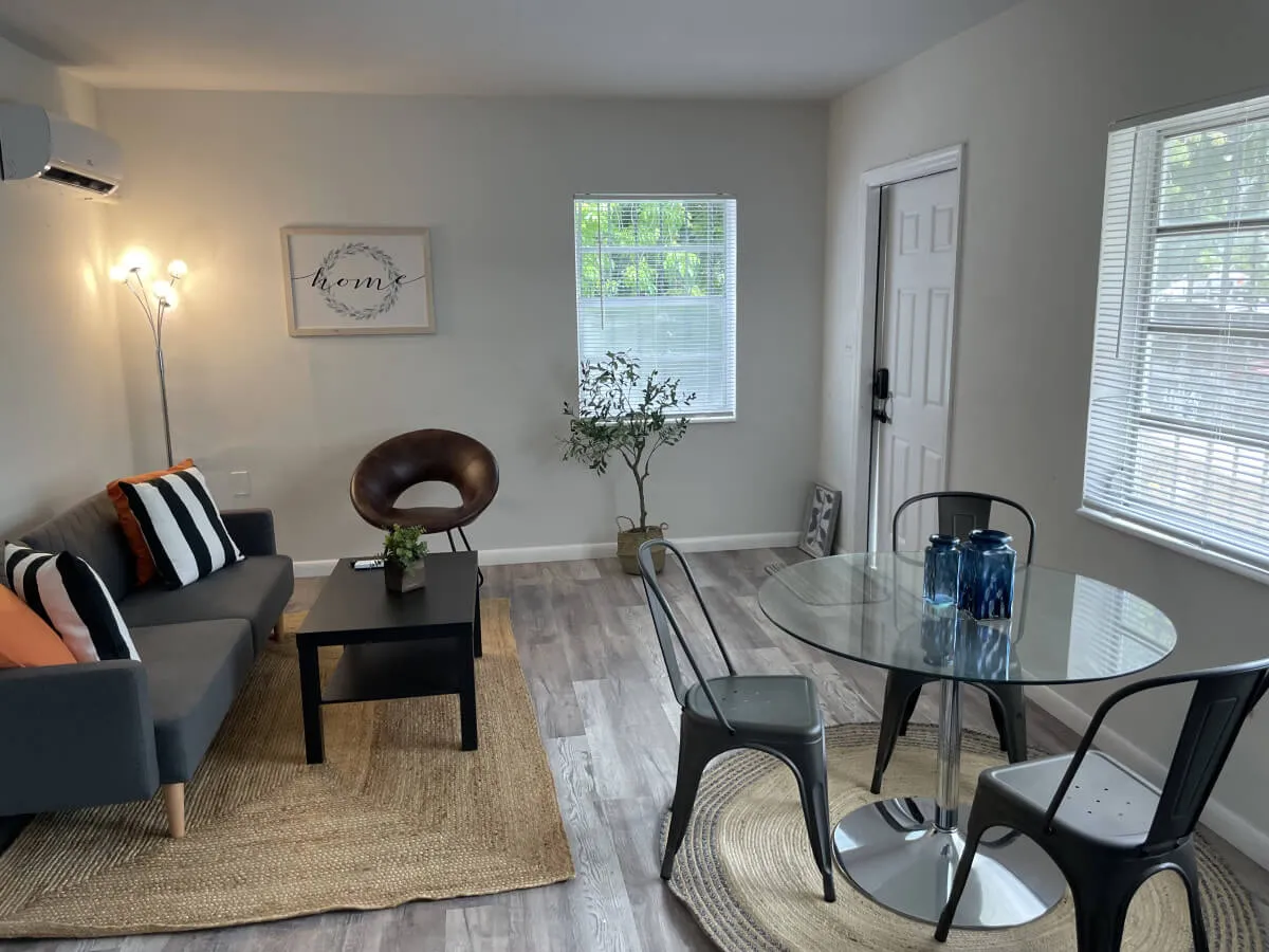 This interior shot shows a bright, comfortable living area with a sofa adorned with colorful cushions, a glass dining table, a decorative plant, and framed artwork featuring the word 'home.' The setup feels welcoming and thoughtfully designed, echoing the entryway’s ambiance mentioned in the section.
