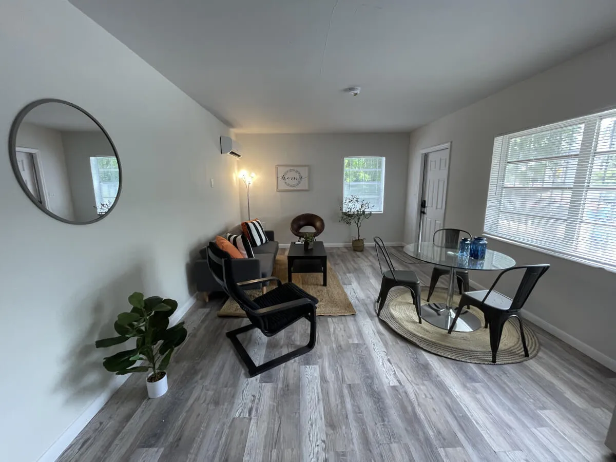 The image showcases a cozy living room featuring a cushioned sofa with throw pillows, a glass dining table set for casual meals, an indoor plant adding a fresh touch, stylish wall art, and a mirrored wall that enhances the sense of space and brightness. Sunlight streams into the room, making it feel airy and welcoming—the ideal spot for relaxing, socializing, or catching up on work.