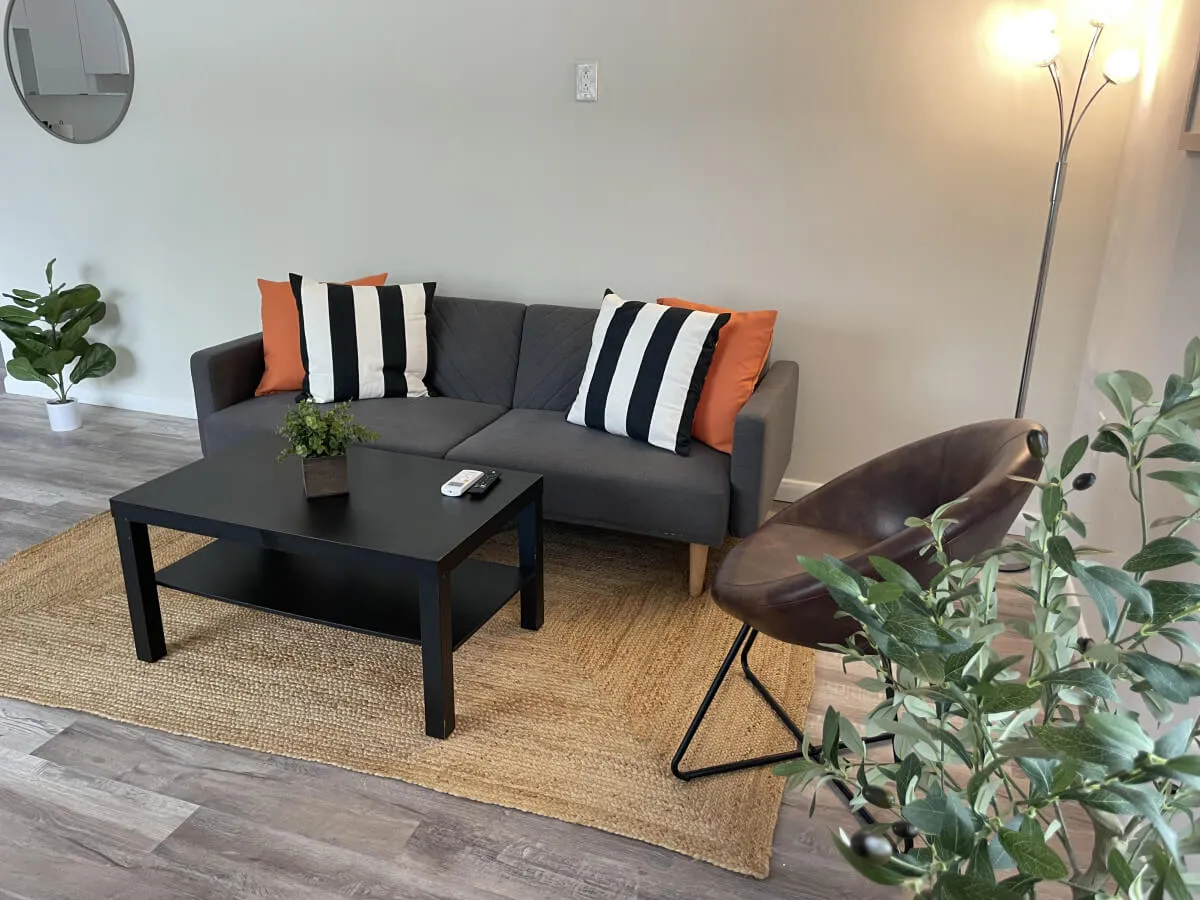 A welcoming living room featuring a comfortable sofa, coffee table, accent chair, and a vibrant green plant. The space is bathed in natural light, offering an inviting atmosphere ideal for relaxing, reading, or gathering as a family after a day of exploration.