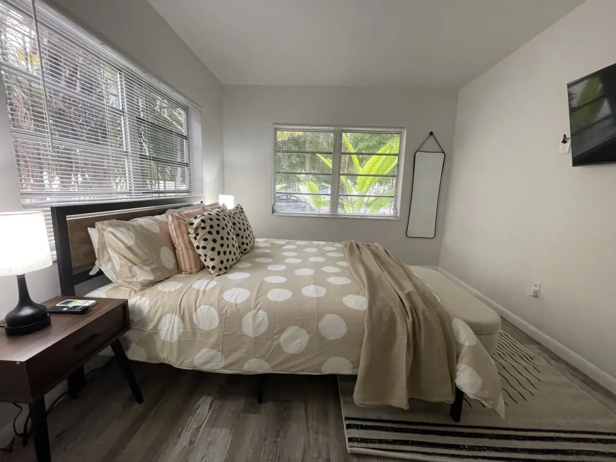 A tranquil bedroom scene with a bed dressed in playful polka dot bedding, a nightstand and lamp for nighttime reading, and a large window that fills the space with natural light, showcasing greenery outside.