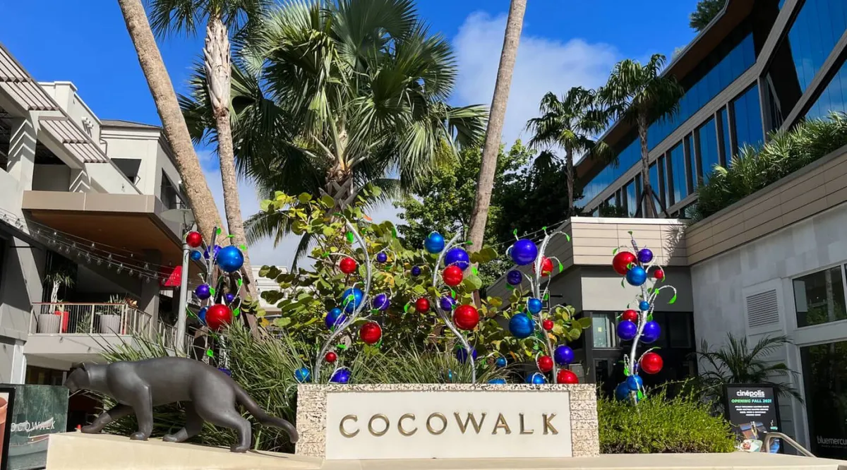 A festive exterior shot of the Cocowalk area, featuring a prominent Cocowalk sign, colorful decorations, a striking panther sculpture, and lush palm trees. The scene is lively and welcoming, hinting at community events and the neighborhood's vibrant atmosphere.