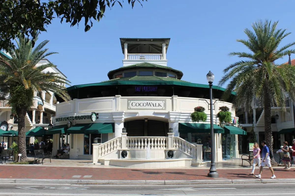 A vibrant scene showing the CocoWalk building surrounded by swaying palm trees, people strolling along the sidewalk, and inviting street lamps. The atmosphere is lively and energetic, capturing the essence of urban Miami with shoppers and diners enjoying the sunshine.