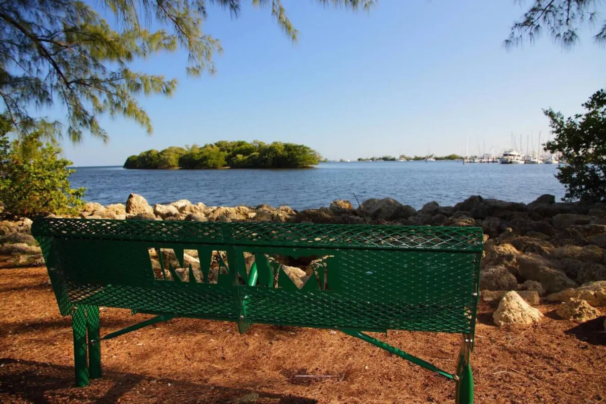 A tranquil waterfront park scene with a green bench featuring a 'Miami' cutout, calm waters, a marina in the background, and a distant island. The setting invites relaxation and offers a glimpse of Miami’s scenic outdoor spaces within easy reach of the city.